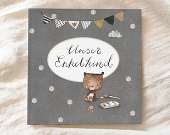 May include: A gray card with a white oval containing the text "Unser Enkelkind" in black cursive writing. A brown bear with a white belly and a paintbrush is sitting on the card with a palette of paints. The card is decorated with a string of flags and silver glitter.