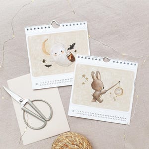 May include: Two white spiral-bound calendars with whimsical illustrations. One features a ghost with a lantern, the other a bunny with a lantern. A pair of scissors and a woven basket are also in the image.