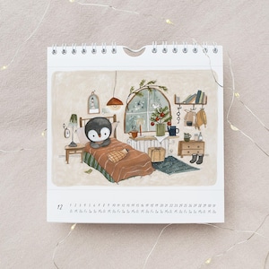 May include: A spiral-bound calendar featuring an illustrated scene of a penguin in bed. The artwork depicts a cozy room with a Christmas tree, books, and a window. The calendar shows the month of December with the days of the month listed.
