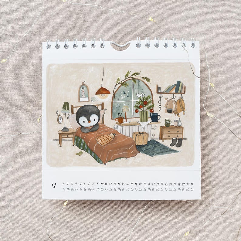 May include: A spiral-bound calendar featuring an illustrated scene of a penguin in bed. The artwork depicts a cozy room with a Christmas tree, books, and a window. The calendar shows the month of December with the days of the month listed.