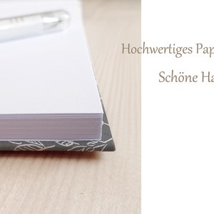 May include: Close-up of a white notebook with a gray and white patterned cover. The notebook is closed and a pen is lying on top of it. The text "Hochwertiges Papier Sch&ouml;ne Haptik" is visible on the right side of the image.