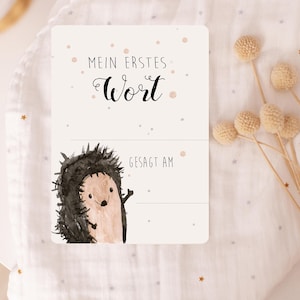 May include: A white card with a watercolor illustration of a hedgehog. The card says "Mein erstes Wort" and has a line for the first word and the date it was said.