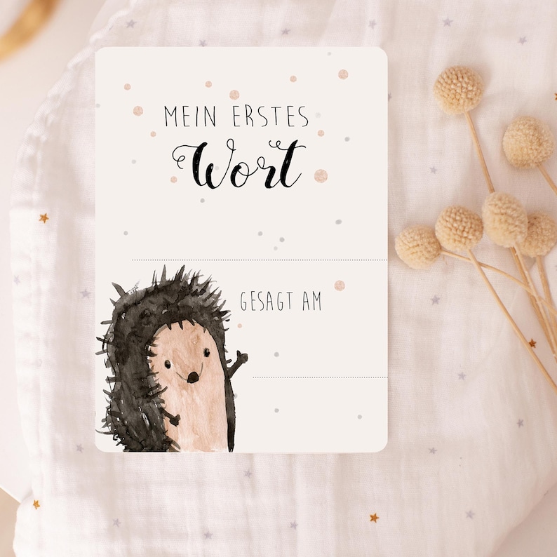 May include: A white card with a watercolor illustration of a hedgehog. The card says "Mein erstes Wort" and has a line for the first word and the date it was said.