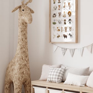 Op de afbeelding: Een ingelijst alfabetposter met illustraties van dieren voor elke letter. De poster hangt aan een witte muur boven een houten bank met opbergbakken. Een geweven giraffenknuffel staat naast de bank.