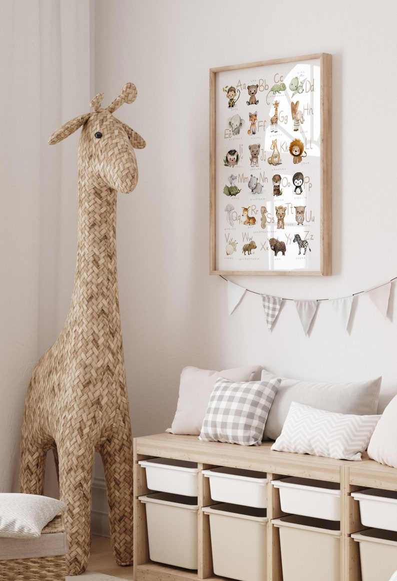 Op de afbeelding: Een ingelijst alfabetposter met illustraties van dieren voor elke letter. De poster hangt aan een witte muur boven een houten bank met opbergbakken. Een geweven giraffenknuffel staat naast de bank.