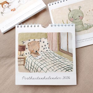 May include: A spiral-bound desk calendar featuring watercolor illustrations. One page shows a teddy bear reading in bed. Other pages display a snowman and a caterpillar. The calendar is labeled "Postkartenkalender 2026".