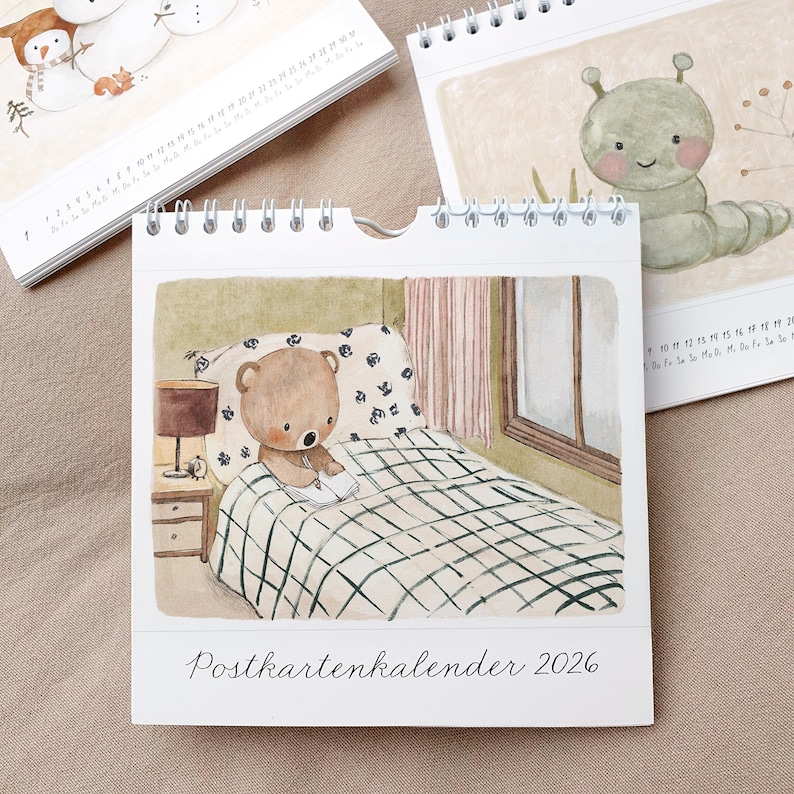 May include: A spiral-bound desk calendar featuring watercolor illustrations. One page shows a teddy bear reading in bed. Other pages display a snowman and a caterpillar. The calendar is labeled "Postkartenkalender 2026".