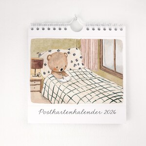 May include: A spiral-bound calendar featuring a watercolor illustration of a teddy bear reading in bed. The bear is on a bed with a checkered blanket and floral pillow. A bedside lamp and the text "Postkartenkalender 2026" are also visible.