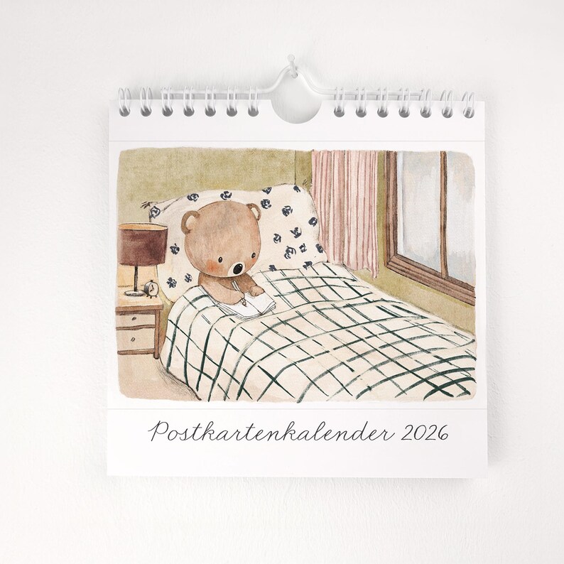 May include: A spiral-bound calendar featuring a watercolor illustration of a teddy bear reading in bed. The bear is on a bed with a checkered blanket and floral pillow. A bedside lamp and the text "Postkartenkalender 2026" are also visible.