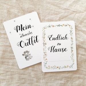 May include: Two white cards with black text. The first card says "Mein allererstes Outfit" with a small illustration of a baby onesie. The second card says "Endlich zu Hause" with a floral border.