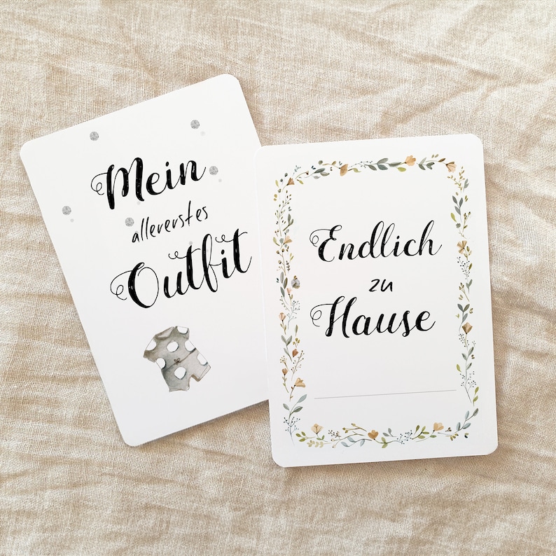 May include: Two white cards with black text. The first card says "Mein allererstes Outfit" with a small illustration of a baby onesie. The second card says "Endlich zu Hause" with a floral border.