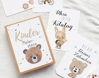 May include: A box of illustrated cards for children, featuring a brown bear with stars and the text "Kinder Meilensteine". The cards are designed for milestones in a child's life, such as their first day of school or losing a tooth.