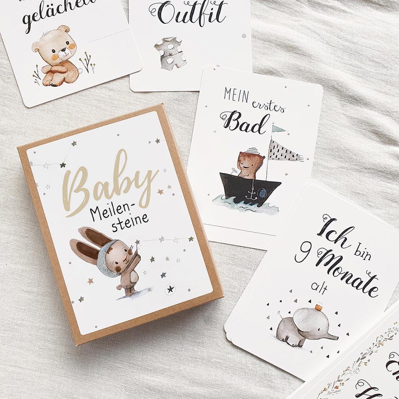 May include: A set of baby milestone cards with illustrations of animals. The cards feature phrases like "Baby Meilensteine", "Outfit", "Mein erstes Bad", and "Ich bin 9 Monate alt".