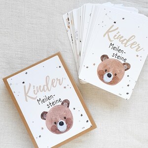 May include: A brown box with a white label that says "Kinder Meilensteine" with a cartoon bear illustration. The box contains a set of cards with the same design and text.