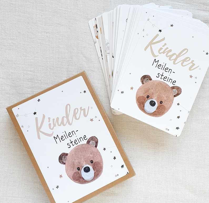 May include: A brown box with a white label that says "Kinder Meilensteine" with a cartoon bear illustration. The box contains a set of cards with the same design and text.