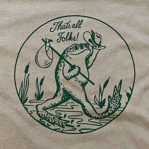 May include: Tan t-shirt with a green circular graphic featuring a frog carrying a bindle and wearing a hat. The text "That's all Folks!" is above the frog. The frog is walking through a marsh with cattails.