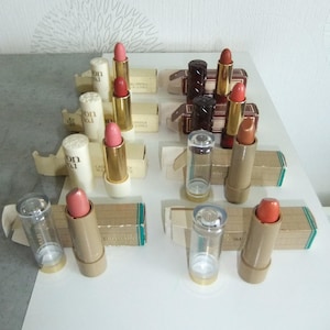 Peut inclure: Une sélection de rouges à lèvres aux teintes rose, rouge et corail, présentés avec leur emballage d'origine. Les rouges à lèvres ont des tubes dorés ou marron et sont disposés sur une surface blanche.