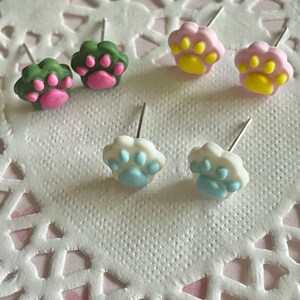 Könnte beinhalten: Sechs Ohrstecker in Form von Pfotenabdrücken. Die Ohrringe sind in drei Farbkombinationen erhältlich: Grün mit rosa Pfoten, Rosa mit gelben Pfoten und Weiß mit blauen Pfoten. Sie liegen auf einer weißen, strukturierten Oberfläche.