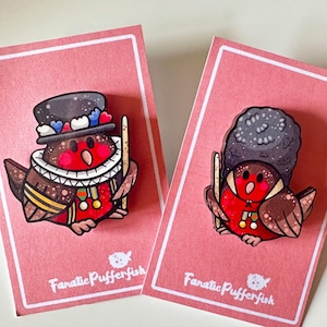 Puede incluir: Dos pines de petirrojos ilustrados en tarjetas rosas. El petirrojo de la izquierda lleva una corona y un sombrero rojo, blanco y azul. El petirrojo de la derecha lleva un sombrero negro y blanco. Ambos petirrojos sostienen una baqueta. Las tarjetas tienen el texto "Fanatic Pufferfish".