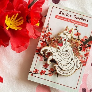 May include: White enamel pin of a nine-tailed fox with gold accents, holding a red bell. The fox is surrounded by red and gold maple leaves. The pin is on a white card with the text "Shrine Dwellers Enamel pins by Fanaticsutferfisch".