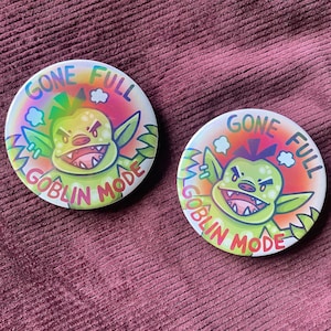 Può includere: Due bottoni rotondi iridescenti con un personaggio di cartone animato di goblin verde e il testo "Gone Full Goblin Mode".