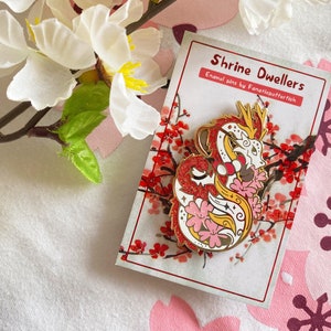 Może przedstawiać: Złota szpilka emaliowana przedstawiająca czerwono-białego smoka z różowymi kwiatami wiśni. Szpilka znajduje się na białej kartce z tekstem "Shrine Dwellers Enamel pins by Fanaticbutterfish".