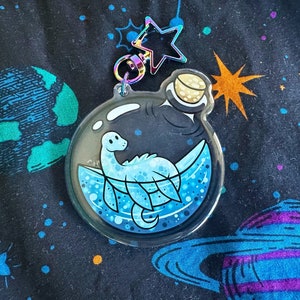 Puede incluir: Un amuleto de botella de vidrio transparente con un tapón de corcho dorado, que contiene una serpiente marina azul y blanca de dibujos animados. El amuleto está sobre un fondo negro con planetas azules y verdes.