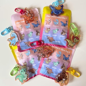 Puede incluir: Varios paquetes de Dino Charm Blind Bag II con colgantes con forma de dinosaurio adjuntos. Los paquetes son amarillos y rosas, con una lista de ilustraciones de dinosaurios. Los colgantes son de varios colores y están unidos a cierres coloridos.
