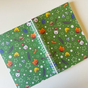 May include: Spiral-bound notebook with a green cover featuring a repeating pattern of cartoon characters, stars, and other fantasy-themed elements. The notebook is open, revealing the white spiral binding and blank pages.