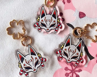 May include: Three gold keychains with clear acrylic charms featuring a white fox mask with red and yellow details. Each charm is attached to a gold chain with a gold flower-shaped ring.