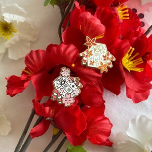 May include: Two gold enamel pins with red and white details. The pins feature a traditional Japanese design with the text "福" and "寿" in gold. The pins are displayed on a background of red and white flowers.