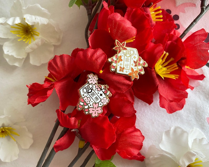 May include: Two gold enamel pins with red and white details. The pins feature a traditional Japanese design with the text "福" and "寿" in gold. The pins are displayed on a background of red and white flowers.