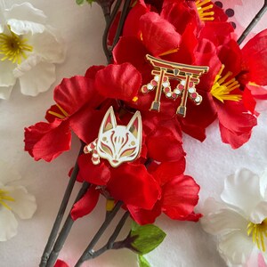 May include: Two gold enamel pins with red and white details. One pin features a red and white Japanese torii gate with white beads. The other pin features a white fox mask with red accents.