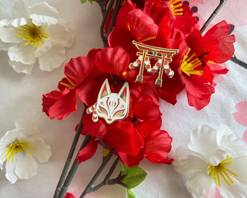 May include: Two gold enamel pins with red and white details. One pin features a red and white Japanese torii gate with white beads. The other pin features a white fox mask with red accents.