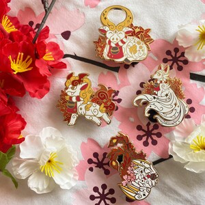 May include: Four enamel pins featuring different mythical creatures. The pins are white with gold accents and red and gold details. The creatures are a fox, a dog, a dragon, and a deer.