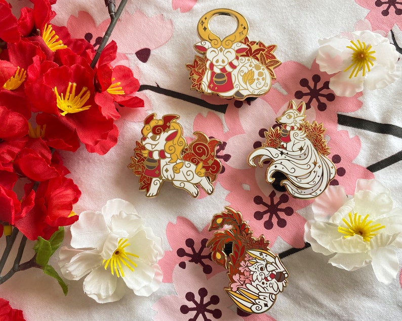 May include: Four enamel pins featuring different mythical creatures. The pins are white with gold accents and red and gold details. The creatures are a fox, a dog, a dragon, and a deer.