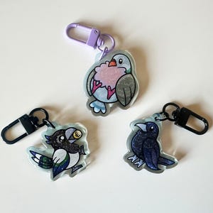 May include: Three colorful bird keychains with glitter accents. The keychains feature a blue and gray bird, a black and white bird, and a blue bird.