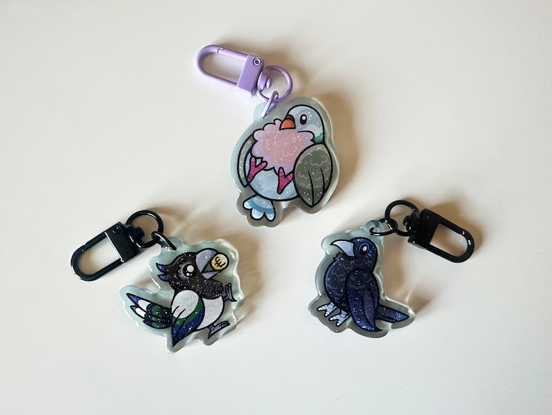 May include: Three colorful bird keychains with glitter accents. The keychains feature a blue and gray bird, a black and white bird, and a blue bird.
