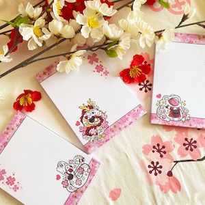 May include: Three pink and white notepad sets with floral designs and illustrations of a dragon, a frog, and a snake. Each notepad has a white background with pink floral accents and a pink border.