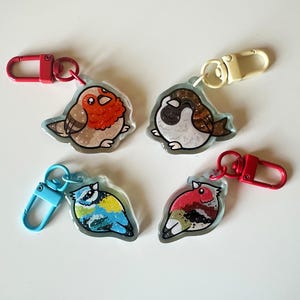 May include: Four colorful bird keychains with glitter accents. Each keychain features a different bird species, including a robin, a sparrow, a bluebird, and a cardinal. The keychains are made of clear resin and have metal clasps.