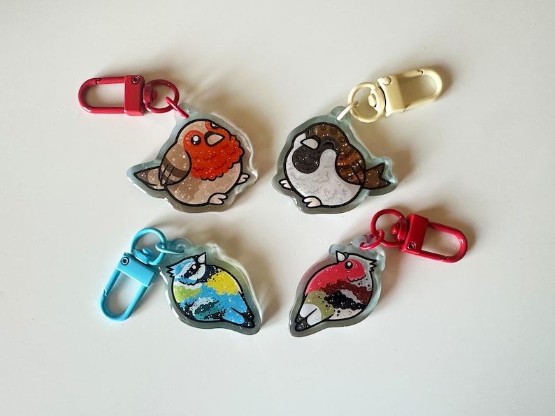 May include: Four colorful bird keychains with glitter accents. Each keychain features a different bird species, including a robin, a sparrow, a bluebird, and a cardinal. The keychains are made of clear resin and have metal clasps.