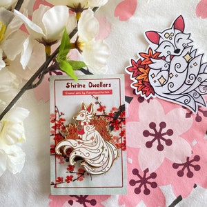May include: A white enamel pin featuring a white fox with red accents and a gold outline. The fox is curled up with its tail forming a circle around its body. The pin is on a white card with red and gold accents. The card has the text "Shrine Dwellers Enamel pins by Fanaticpufftah" printed on it.