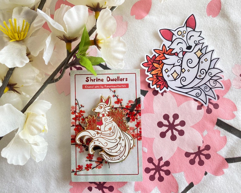 May include: A white enamel pin featuring a white fox with red accents and a gold outline. The fox is curled up with its tail forming a circle around its body. The pin is on a white card with red and gold accents. The card has the text "Shrine Dwellers Enamel pins by Fanaticpufftah" printed on it.