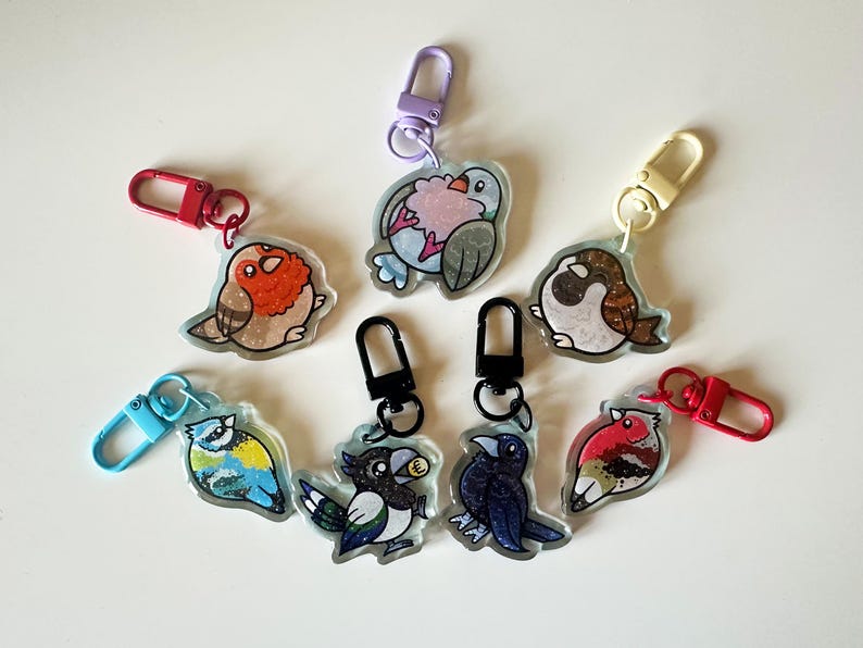 May include: A set of eight colorful bird keychains with glitter accents. Each keychain features a different bird species, including a robin, a blue jay, a cardinal, and a sparrow. The keychains are made of clear acrylic and have metal clasps.