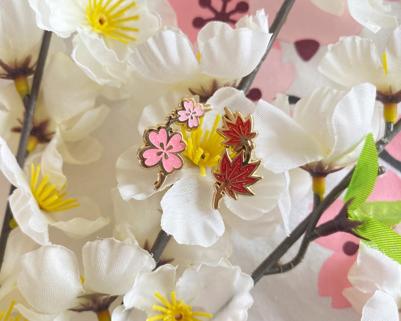 May include: Gold-tone enamel pin featuring a cluster of pink cherry blossoms and red maple leaves. The pin is set against a background of white fabric flowers.