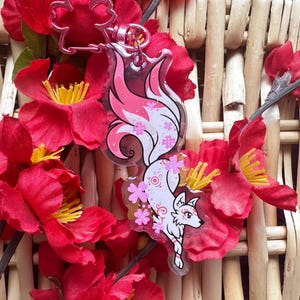 May include: A clear acrylic keychain featuring a stylized white and pink fox with floral accents. The keychain is attached to a silver-colored clasp and is surrounded by vibrant red flowers with yellow centers, set against a woven background.