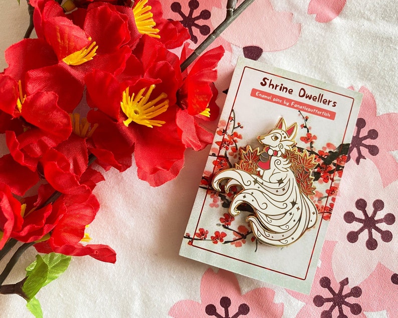 May include: White enamel pin of a nine-tailed fox with gold accents, holding a red bell. The fox is surrounded by red and gold maple leaves. The pin is on a white card with the text "Shrine Dwellers Enamel pins by Fanaticsutferfisch".