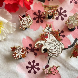 May include: A set of nine enamel pins featuring Japanese-inspired designs. The pins include a fox with a nine-tailed, a kitsune mask, a torii gate, a red and white envelope with the Japanese characters "lucky charm", a red and white envelope with the Japanese characters "good luck", and a cluster of red maple leaves. The pins are all gold-toned with red and white accents.
