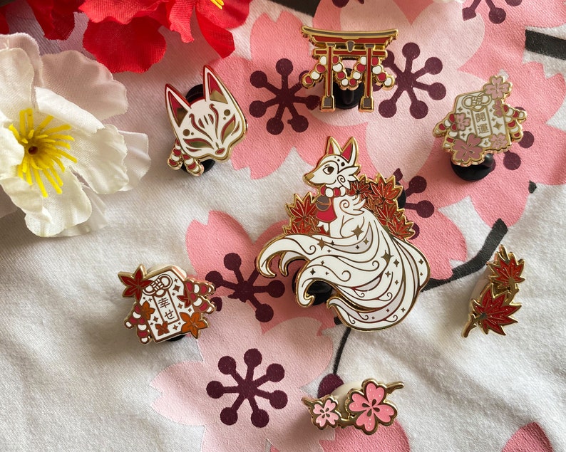 May include: A set of nine enamel pins featuring Japanese-inspired designs. The pins include a fox with a nine-tailed, a kitsune mask, a torii gate, a red and white envelope with the Japanese characters "lucky charm", a red and white envelope with the Japanese characters "good luck", and a cluster of red maple leaves. The pins are all gold-toned with red and white accents.