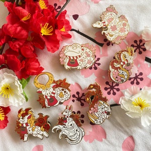 May include: A set of seven enamel pins featuring various mythical creatures, each with a unique design. The pins are gold and white with red and pink accents. The creatures include a frog, a fox, a dragon, a phoenix, a kitsune, a tanuki, and a kappa.